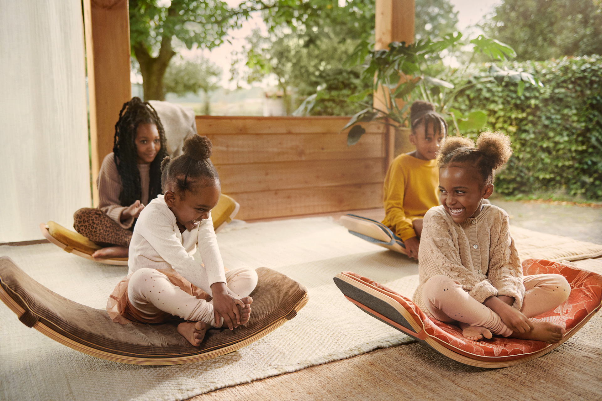 Vier meisjes zitten verdeeld over de kamer. Onder hen ligt een licht kleurig tapijt, elks van hen zit op een hobbel. De rest van de kamer heeft hout kleuren en planten op de achtergrond Vier meisjes zitten verdeeld over de kamer. Onder hen ligt een licht kleurig tapijt, elks van hen zit op een hobbel. De rest van de kamer heeft hout kleuren en planten op de achtergrond